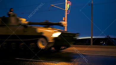 A convoy of military equipment rides through the city at night with headlights on