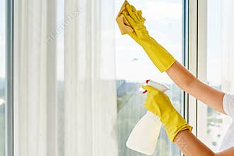 Close up of woman in yellow rubber gloves cleaning window with cleanser spray and yellow rag at home or office, copy space.