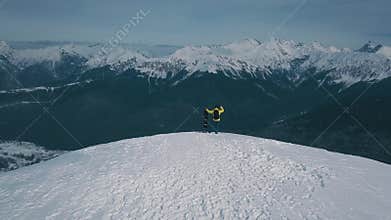 Aerial shot flying over winner male snowboarder admiring mountain landscape from peak of rock