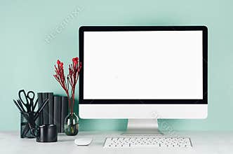 Simple office interior with blank computer monitor, black stationery, books, red flowers, keyboard, coffee cup in green mint.