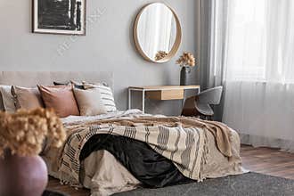 Elegant round mirror in wooden frame above fancy console table with flowers in vase in trendy bedroom interior with beige vase