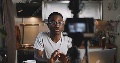 Happy young successful black vlogger man filming video for his online blog with camera at home kitchen slow motion.