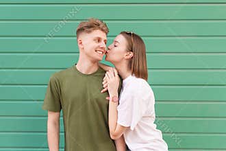 Young lovers are a couple of teens standing on the background of a green wall, the boy smiles, the girl kisses his cheek. Young