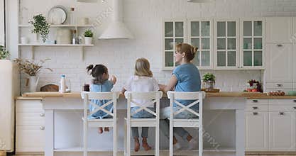 Mom and daughters sit on chairs in kitchen, rear view