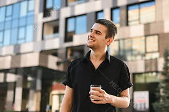 Happy young man in a stylish black shirt stands against the backdrop of modern architecture with a paper cup of coffee in his hand