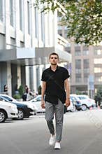Full length portrait of a young man in stylish casual clothes, wearing a black shirt and gray pants, walking down the city street