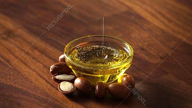 Argan oil pouring into a glass bowl with argan seeds on a wooden background in sunlight. Argan oil based cosmetics concept. Copy