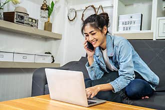 Asian young business woman is talking on cell phone and smiling with laptop computer at home office, work from home