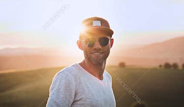 Smiling at camera handsome cheerful man with 3-Day Stubble Beard, baseball cap and fancy blue sunglasses with bright sunset sun