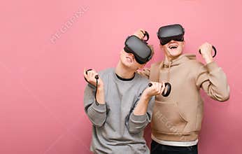 Two cheerful friends standing on a pink background with VR helmets on heads and controllers in their hands and playing video games