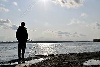 The silhouette of a fisherman, with a spinning rod in hand, fishes on the river, against the blue of water and the sky