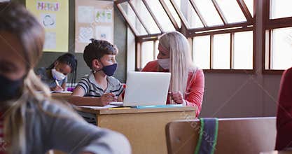 Female teacher and boy wearing face mask using laptop in class at school