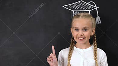 Schoolgirl pointing finger at blackboard, academic cap painted above head