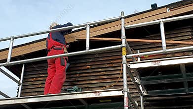 Worker on scaffolding installing new wooden planks on house roof eaves