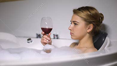 Depressed girl lying in bath with foam bubbles drinking wine, boyfriend break up