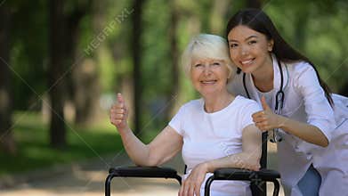 Nurse and elderly woman in wheelchair smiling at camera and showing thumbs-up