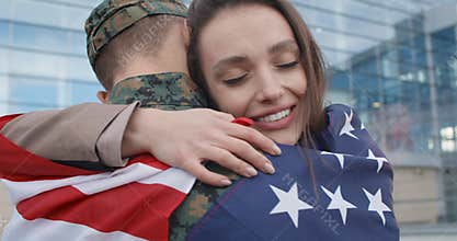 Happy girl embracing military boyfriend covered with american flag.