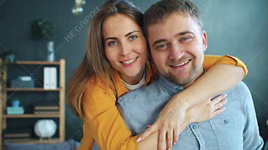 Portrait of happy young woman kissing and hugging husband at home looking at camera