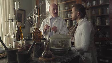 Concentrated Caucasian young man taking notes with feather pen. His colleague pharmacist showing at ancient chemical