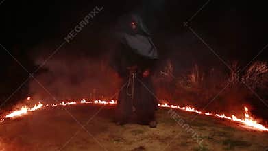 A man in a black monk's robe stands at night in the middle of a ring of fire, a magical ancient rite