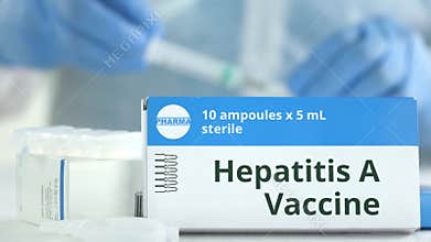 Box with hepatitis A vaccine on the table against blurred lab assistant or doctor. Fictional phaceutical logo
