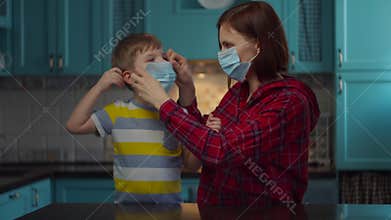Young 30s mother in medical mask helps kid to wear medical mask on children face at home. Woman and boy in masks looking