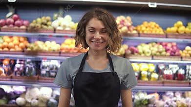Portrait of attractive young saleswoman in black apron standing in supermarket with shelves of fruits on background