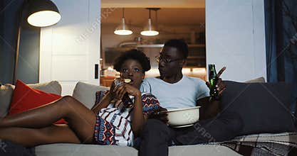 Happy young African positive friends couple watching TV at home on the couch, eating snacks and laughing slow motion.