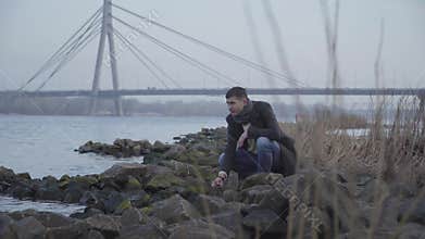 Thoughtful Caucasian young man sitting on riverbank and throwing rocks in water. Portrait of sad handsome guy in