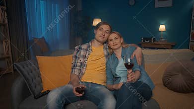 A young couple sits hugging on the couch with glasses of red wine and watching TV. A man and a woman are watching a