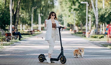 Beautiful young woman in white suit and sunglasses standing with her electric scooter and Corgi Dog in city parkland and drinking