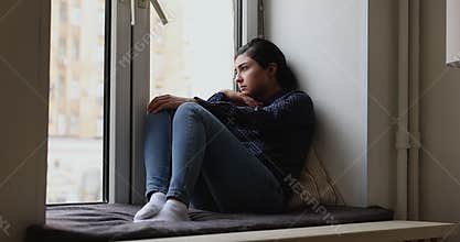 Depressed passive young Indian woman sitting on windowsill