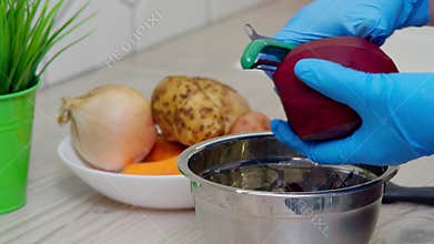 Closeup cropped hand in blue gloves peeling beetroot fast with special blade peeler above metal pan near vegetable plate