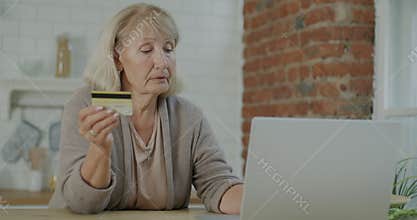 Elderly woman paying with credit card using laptop shopping online indoors in kitchen in apartment