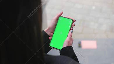 Close up female hands holding a vertical mobile phone with green screen against background of urban city. Chroma key