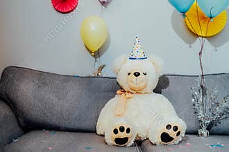 Big Teddy bear with balloons in party cap sitting on the gray sofa in decorated room. Birthday or festive holiday