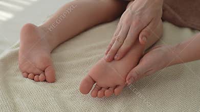 Close-up hands of unrecognizable female pediatric masseuse massaging heel foot of five year old boy client lying on
