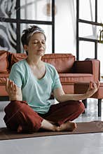 A middle aged lady is practicing yoga at home. A woman with closed eyes sits relaxed on a mat on the floor in the lotus