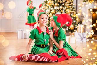 Two cheerful happy girls-friends in costumes of Santa`s elves on the background of a Christmas tree and other children