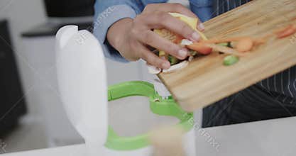 Hands of biracial woman in apron composting vegetable waste in kitchen, slow motion