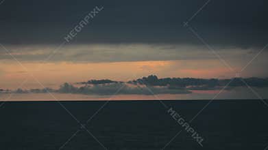 Scenic view of a sea with calm waves and remote orange shades on a dark, cloudy sky