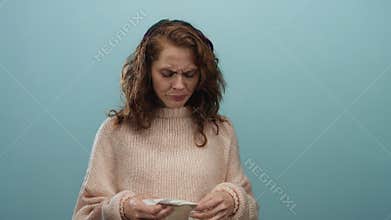 Young woman with curly hair holding a tissue against a blue wall, wearing a cozy sweater, expresses confusion and frustration in