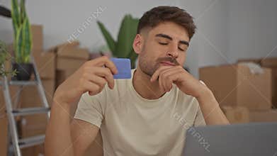 Young hispanic man holds credit card and rests chin on hand in building; thoughtful budget planning