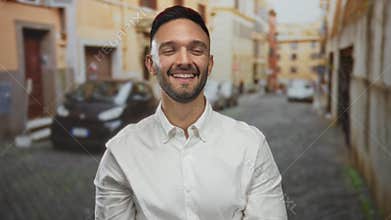 Smiling hispanic man standing confidently with crossed arms in an urban street of an old town city wearing a white shirt outdoors