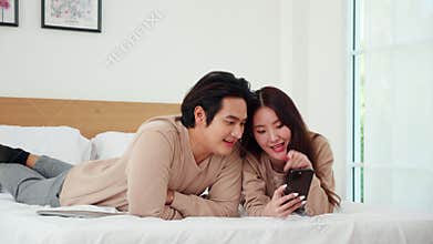 Asian couple lying on bed sharing smartphone together on bed in bedroom at home, digital bonding, relax leisure and relationship,