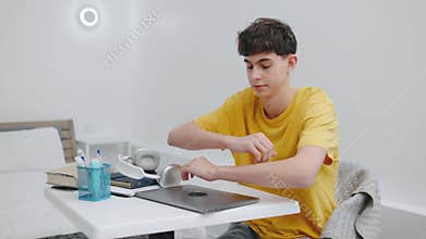 A tired teenager removes his headphones, closes his laptop, and falls asleep on the desk with his head resting on the