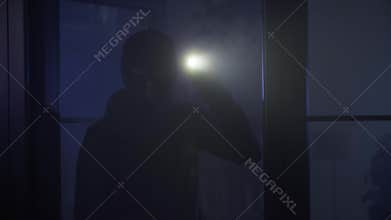 crime and criminality, an adult man in black clothes and a balaclava with flashlight in his hands enters a room and