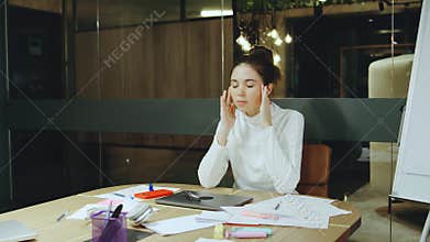 A woman in a well-lit, modern office closes her laptop, removes her glasses, and holds her head in exhaustion after