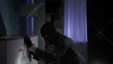 danger, adult man in balaclava and gloves rummages through things in search of money and valuables in apartment