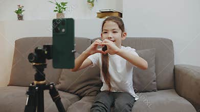 Young girl making a heart shape while video chatting at home, expressing love and joy in a cozy living space during the